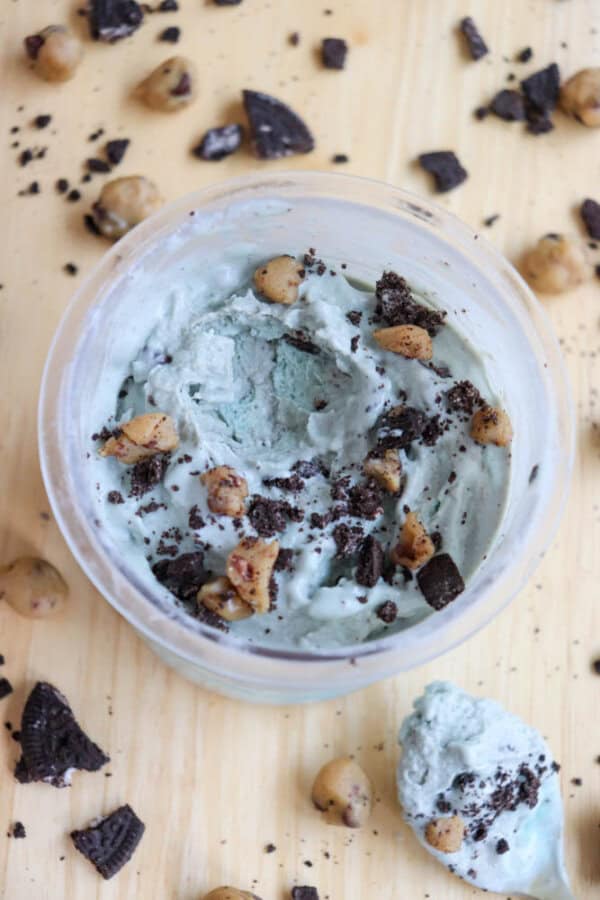 A bowl of pale blue ice cream mixed with chocolate chunks and cookie dough pieces, set on a light wooden surface. Some chocolate and cookie dough pieces are scattered around the bowl, with a spoonful of ice cream beside it.