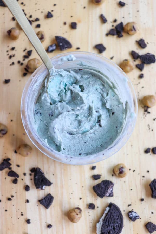 A jar filled with light blue, creamy ice cream sprinkled with chocolate bits. A spoon is dipped into the dip. Around the jar are scattered pieces of chocolate cookies and small round chocolate chip cookie dough bites on a wooden surface.