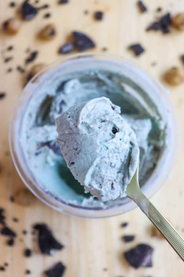 A close-up of a spoon holding a scoop of cookie monster ice cream above an open container. The creamy ice cream is pale green with chocolate chunks. Chocolate pieces and nuts are scattered on a wooden surface in the background.