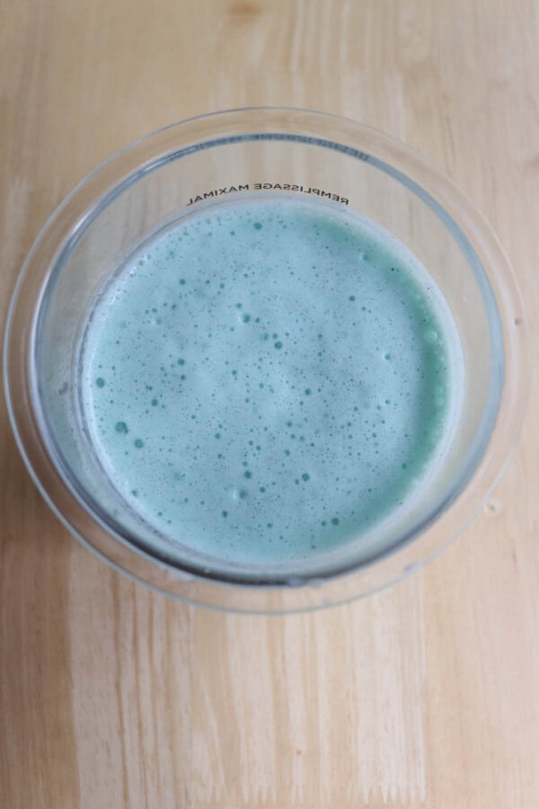A top-down view of a clear glass filled with a frothy, light blue liquid. The glass is placed on a light wooden surface.