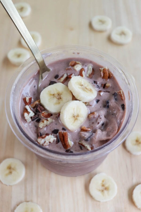 A cup of chocolate smoothie topped with banana slices, pecans, and chocolate chips. A spoon is placed inside the cup, and banana slices are scattered on the wooden surface around the cup.