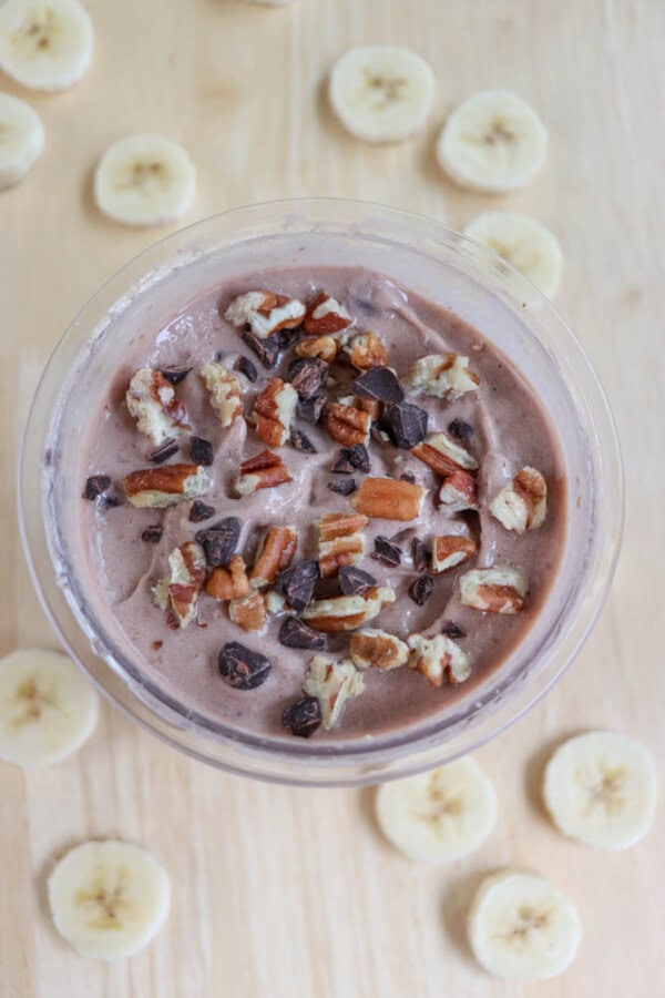 A bowl of chocolate smoothie topped with chopped nuts and dark chocolate chunks sits on a wooden surface. Slices of banana are scattered around the bowl.