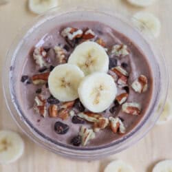 A bowl of chocolate ice cream topped with banana slices, pecans, and chocolate chips. Surrounding the bowl are additional banana slices scattered on a beige surface.