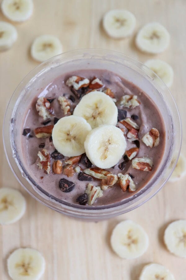 A bowl of chocolate ice cream topped with banana slices, pecans, and chocolate chips. Surrounding the bowl are additional banana slices scattered on a beige surface.
