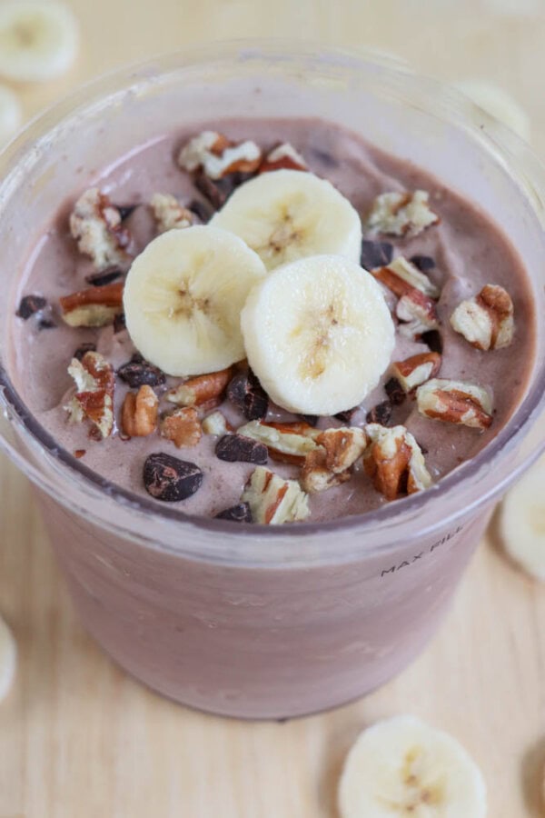 A smoothie bowl topped with banana slices, chopped nuts, and chocolate chips in a clear container. The bowl is placed on a wooden surface scattered with more banana slices.