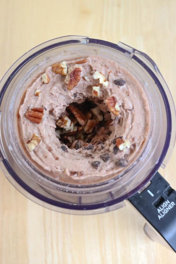 Top view of a food processor containing a creamy mixture of chocolate chip ice cream blended with pecans. The mix is swirled, and the pecans and chocolate chips are visible throughout. The background is a light wooden surface.