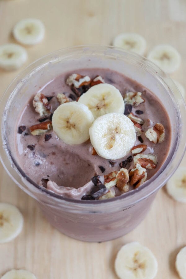 A chocolate smoothie topped with banana slices, chopped nuts, and cacao nibs in a clear plastic cup. The cup is surrounded by additional banana slices on a wooden surface.