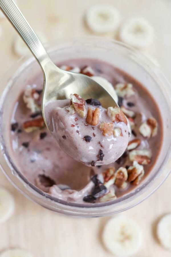 A spoonful of chocolate banana smoothie topped with chopped pecans and cacao nibs, lifted above a glass filled with the same smoothie. Slices of banana are scattered on the light wooden surface below.