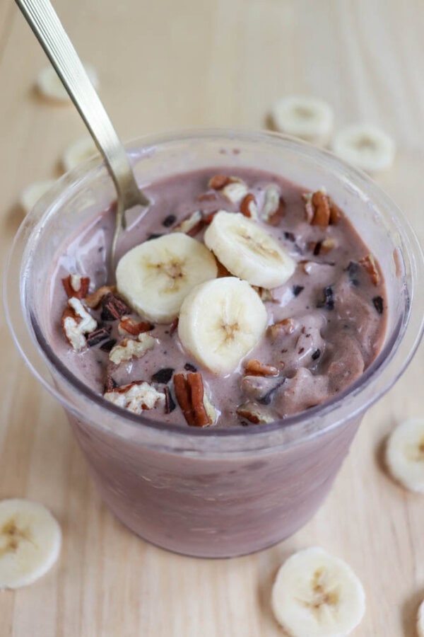 A cup of chocolate oatmeal topped with banana slices, pecans, and chocolate shavings. A spoon is placed inside the cup. Sliced bananas are scattered on the wooden surface around the cup.