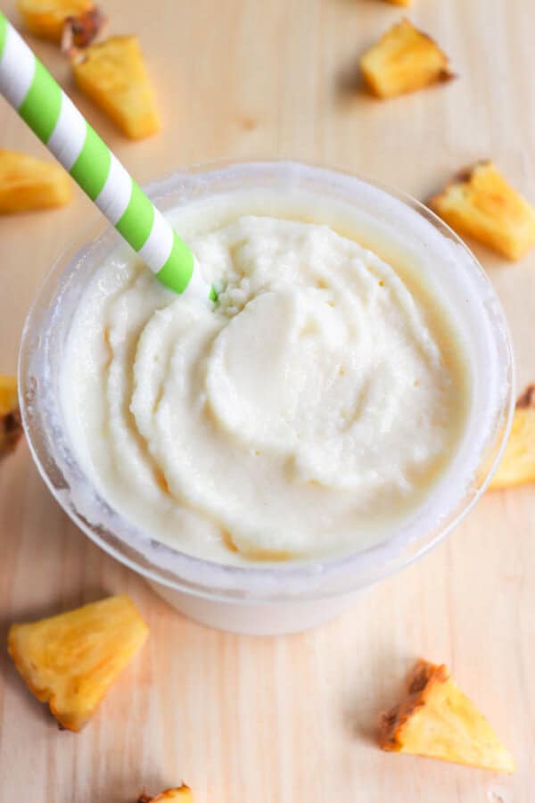A creamy, frothy pineapple dole whip in a clear cup with a green and white striped paper straw. The smoothie is surrounded by small pineapple chunks on a light wooden surface.