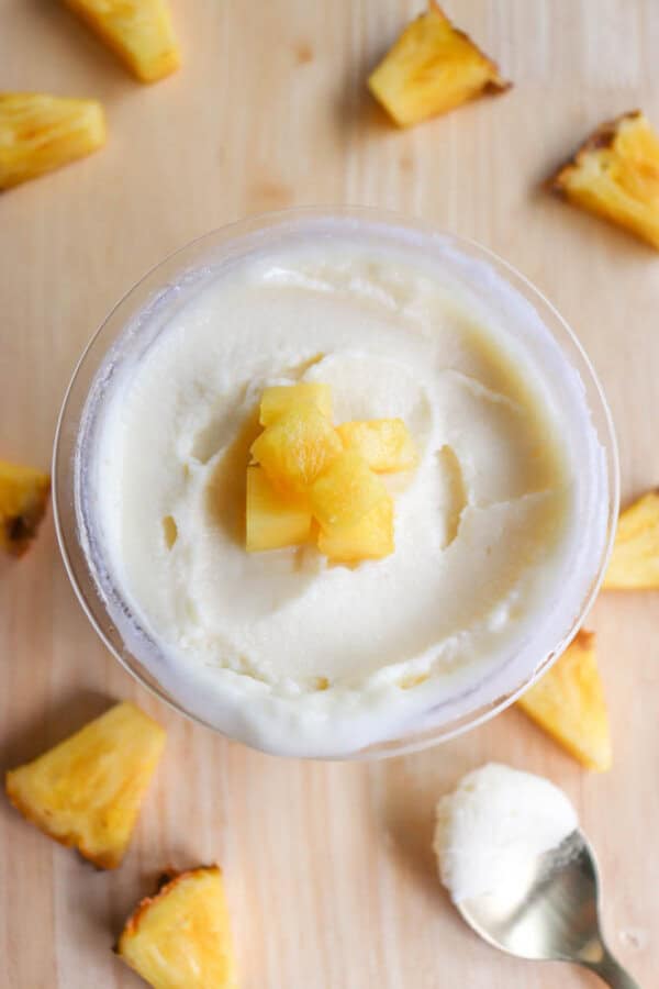 A bowl of creamy, white frozen yogurt topped with small pineapple chunks sits on a wooden surface. Pineapple pieces are scattered around, and a silver spoon with a dollop of yogurt is placed nearby.