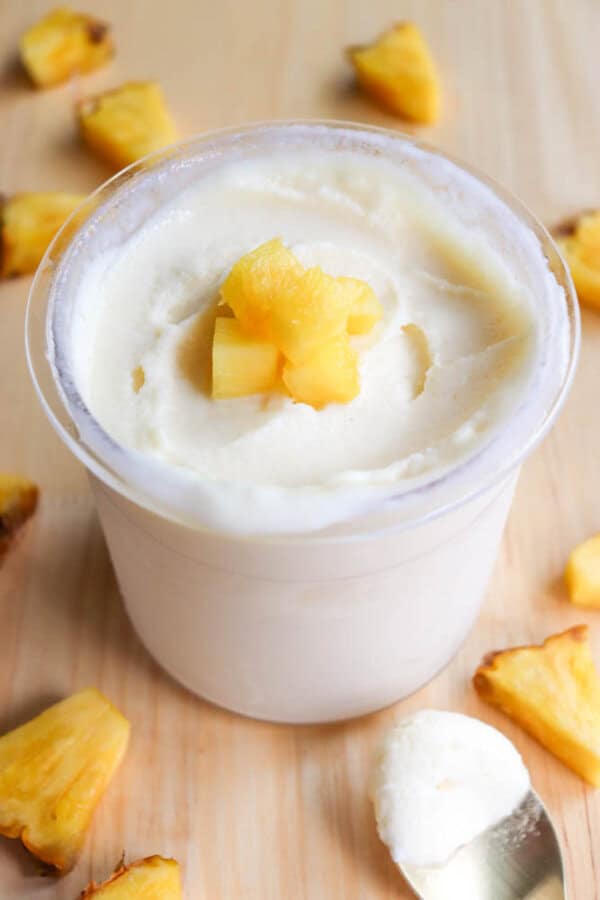 A creamy dole whip topped with diced pineapple chunks is in a clear cup. More pineapple pieces are scattered around the cup on a wooden surface. A spoon with a scoop of the dessert rests nearby.