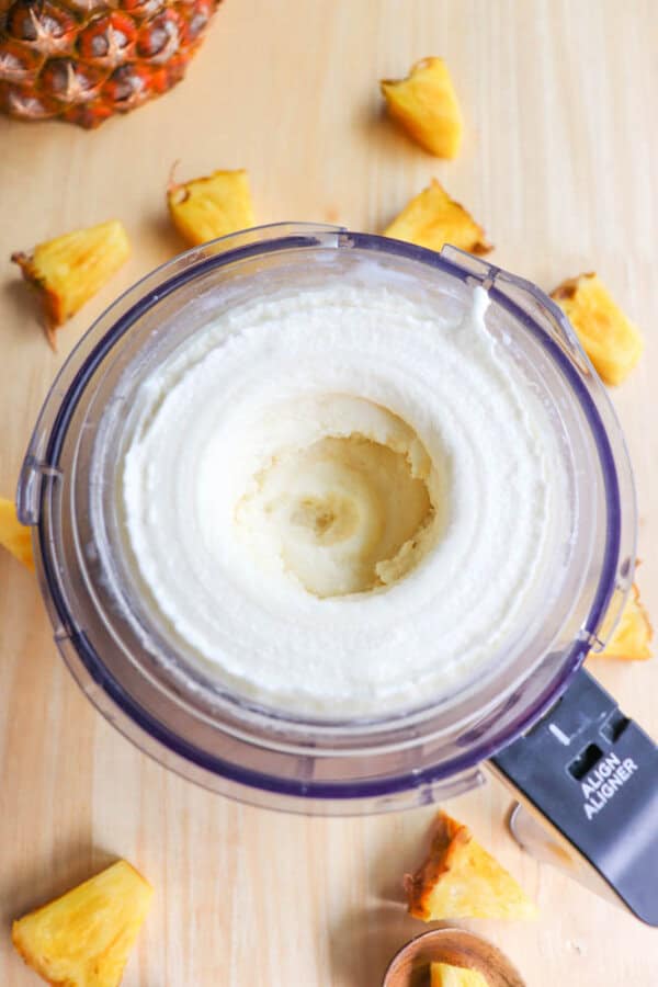 A ninja processor filled with creamy, freshly made pineapple Dole Whip sits on a wooden surface. Pineapple chunks are scattered around the processor, with part of a pineapple visible in the top left corner.