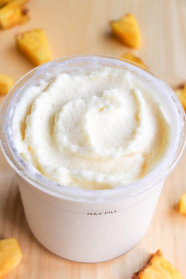 A creamy, white frozen dessert fills a clear cup labeled MAX FILL. The dessert has a swirl pattern on top. Small chunks of pineapple are scattered around the cup on a wooden surface.