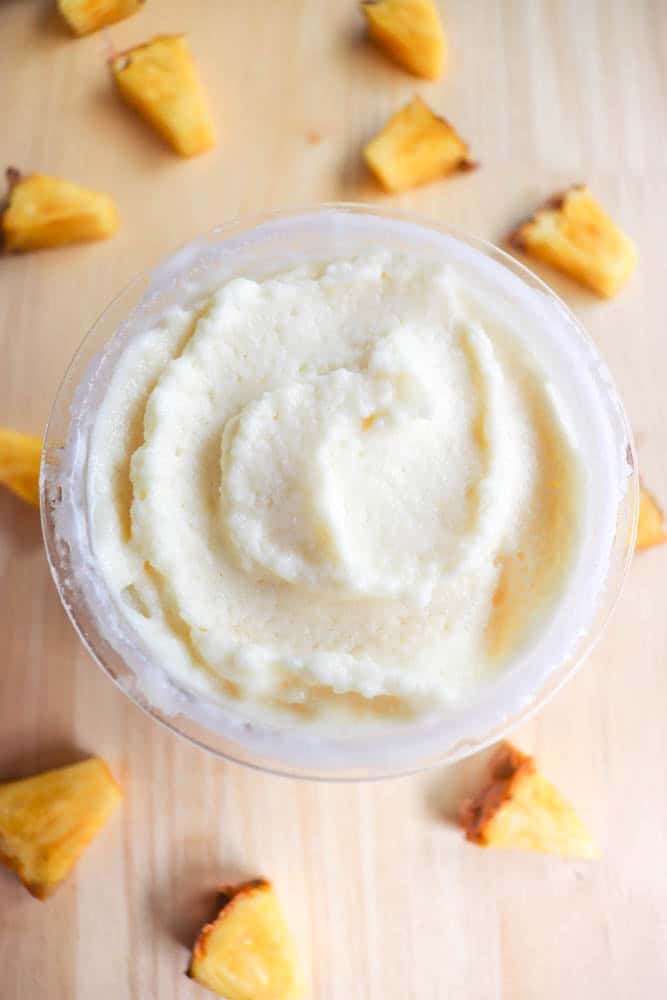 A creamy dessert in a cup, surrounded by small pineapple chunks on a wooden surface. The dessert has a smooth, swirled texture on top, with the pineapple pieces arranged in a scattered pattern around it.