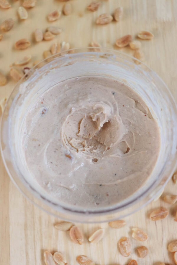 A jar of creamy peanut butter with a smooth texture is placed on a wooden surface. Scattered roasted peanuts surround the jar, enhancing the rustic appearance.