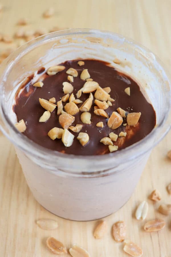A cup filled with chocolate yogurt topped with crushed peanuts, set on a light wooden table with scattered peanuts around. The yogurt displays a smooth chocolate layer on top.