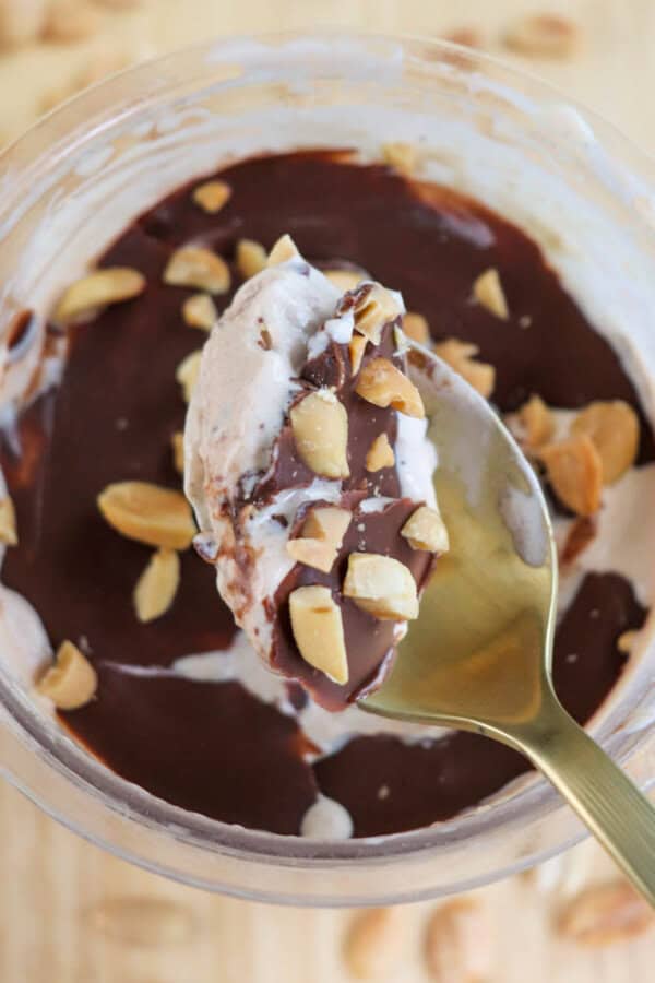 A spoonful of creamy dessert topped with chocolate sauce and chopped peanuts is held above a bowl. The dessert in the bowl features layers of chocolate and nuts, with a wooden background scattered with peanuts.