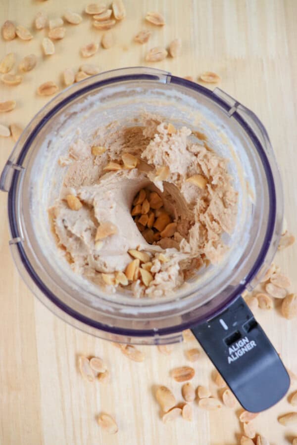 A top view of creamy peanut butter being made in a food processor, surrounded by scattered whole peanuts on a wooden surface. The peanut butter is partially blended, with visible peanut chunks.