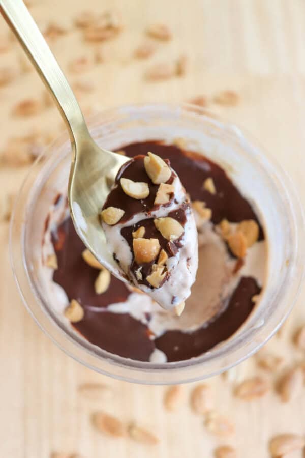 A spoon holding a scoop of ice cream topped with chopped peanuts and chocolate sauce is above a cup filled with the same dessert. Peanut pieces are scattered on the wooden surface around the cup.
