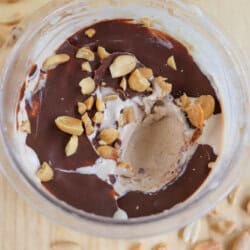 A bowl of chocolate-topped ice cream with a scoop missing, sprinkled with peanuts. Surrounding the bowl are additional scattered peanuts on a wooden surface.