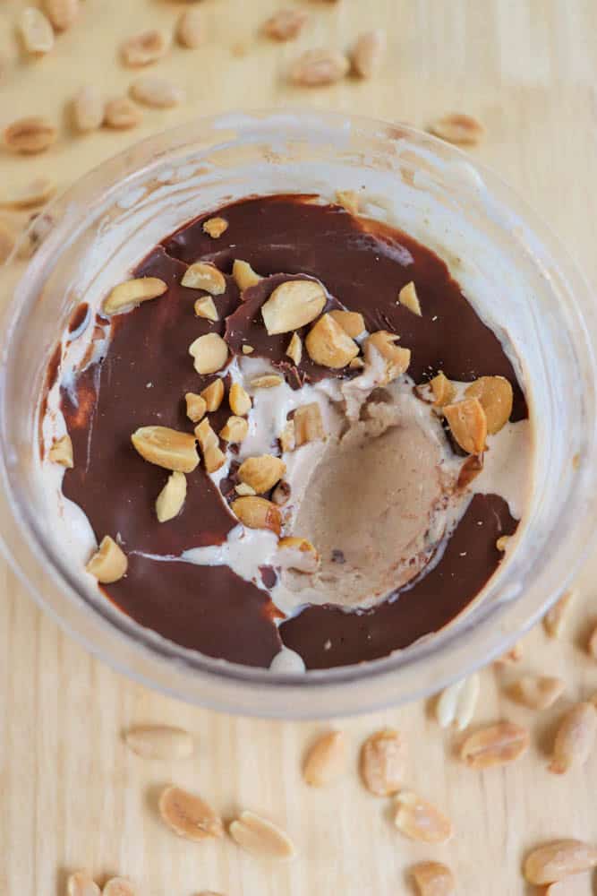 A bowl of chocolate-topped ice cream with a scoop missing, sprinkled with peanuts. Surrounding the bowl are additional scattered peanuts on a wooden surface.