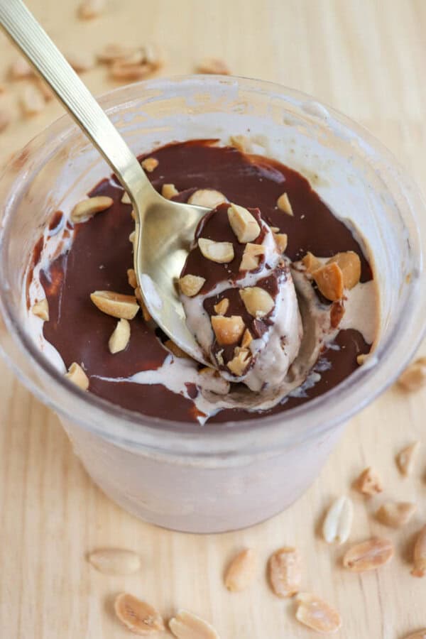A cup of ice cream topped with a layer of chocolate sauce and chopped peanuts. A spoon is scooping a bite from the dish. Peanuts are scattered around the cup on a light wooden surface.