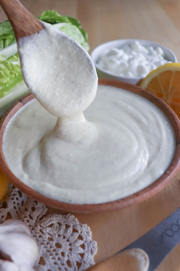 A wooden spoon scoops creamy cottage cheese dressing from a bowl on the table, which also holds garlic cloves, a doily, a knife, lettuce, and a lemon. In the background is a small bowl of Caesar salad dressing.