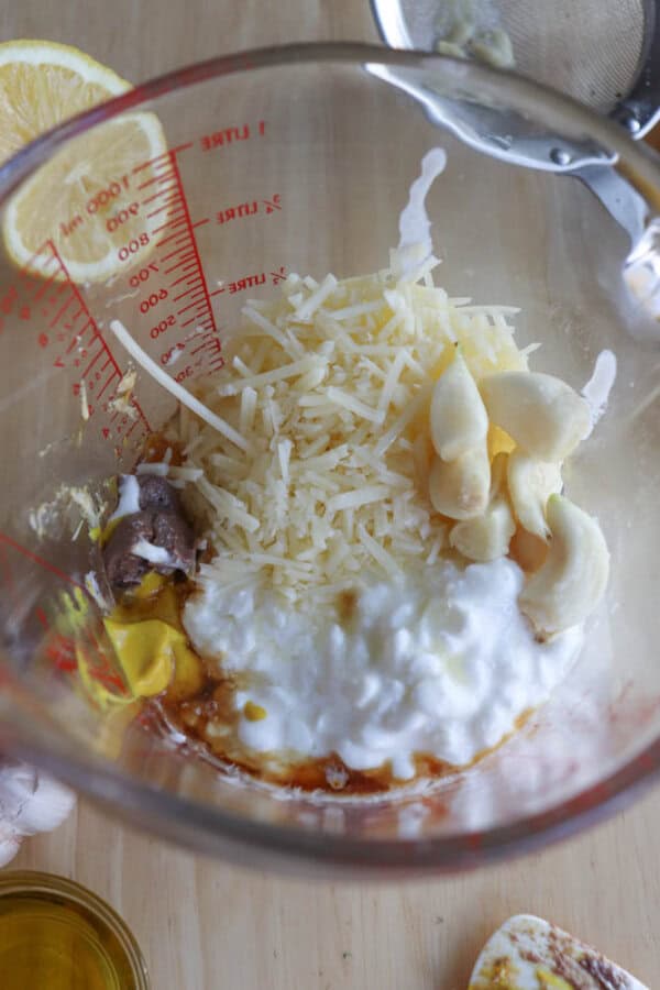 A glass measuring cup holds a medley of ingredients: grated cheese, garlic cloves, anchovies, creamy cottage cheese Caesar salad dressing, an egg yolk, and a brown liquid. Nearby on the wooden surface lie half a lemon and a small bowl of olive oil.