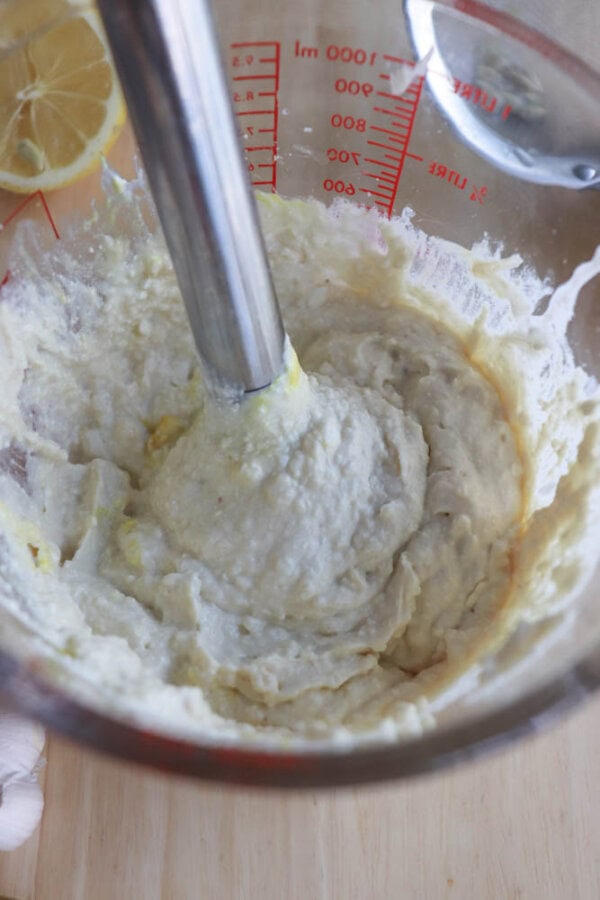 Close-up of an immersion blender mixing a creamy white garlic sauce, like a rich cottage cheese Caesar salad dressing, in a measuring cup. Lemon slices and garlic cloves are visible in the background on a wooden surface.