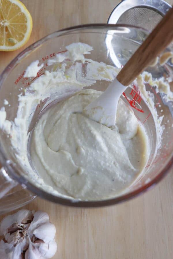 A glass bowl on a wooden surface holds creamy hummus being blended with a spatula. Nearby are a lemon half, a garlic bulb, and a metal strainer, suggesting the artful preparation of an exquisite caesar salad dressing.