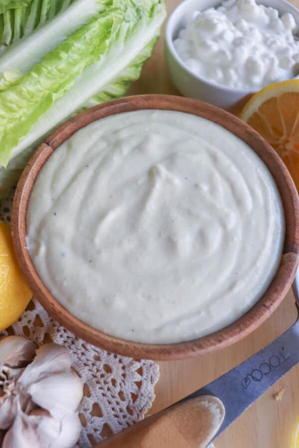 A wooden bowl of creamy Caesar salad dressing sits on a table, surrounded by fresh lettuce, garlic cloves, lemon, and a small bowl of cottage cheese. Nearby, a knife with a wooden handle awaits the next culinary task.