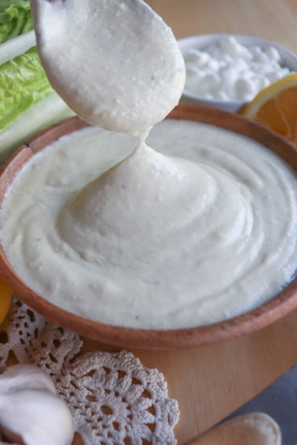 A spoon drizzles creamy garlic sauce into a wooden bowl, creating the perfect base for a cottage cheese Caesar salad dressing. The scene is set with lettuce, lemon slices, a doily, garlic cloves, and crumbled cheese on a rustic wooden surface.