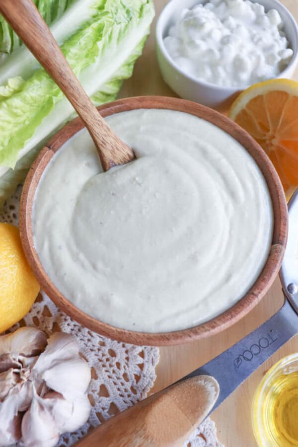 A wooden bowl of creamy Caesar salad dressing with a wooden spoon. Surrounding items include lettuce, a halved lemon, garlic, and a knife with a wooden handle. A small bowl of crumbled cheese on the lace cloth completes this delightful cottage cheese-inspired scene.