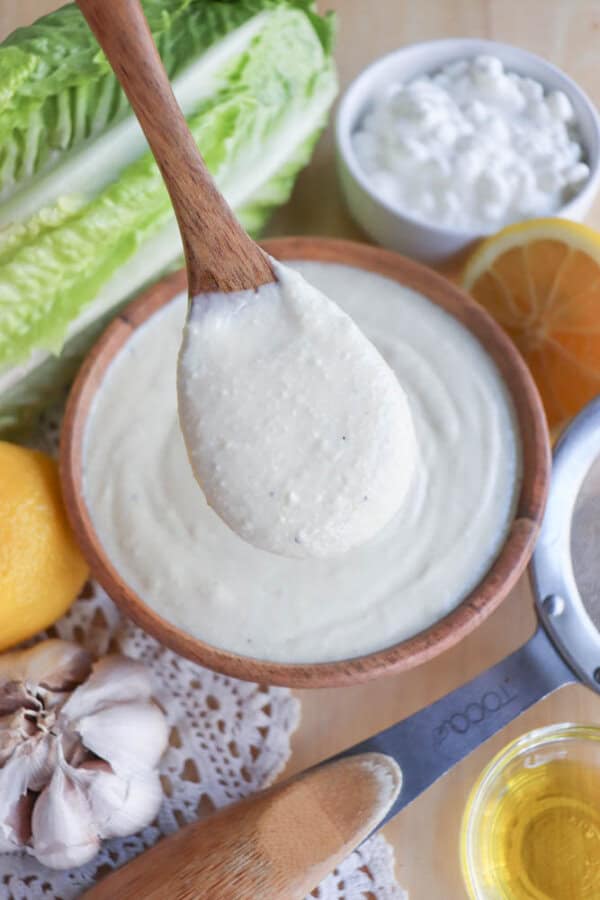 A wooden spoon drizzles creamy cottage cheese Caesar salad dressing over a bowl, surrounded by romaine lettuce, lemon, garlic, and olive oil on a lace cloth background.