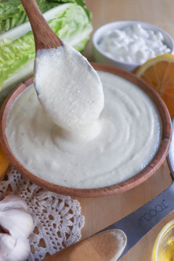 A wooden spoon lifts creamy Caesar salad dressing from a wooden bowl. Surrounding items include fresh lettuce, a small bowl of cheese, a doily, garlic cloves, a lemon slice, and a partially visible cheese slicer.