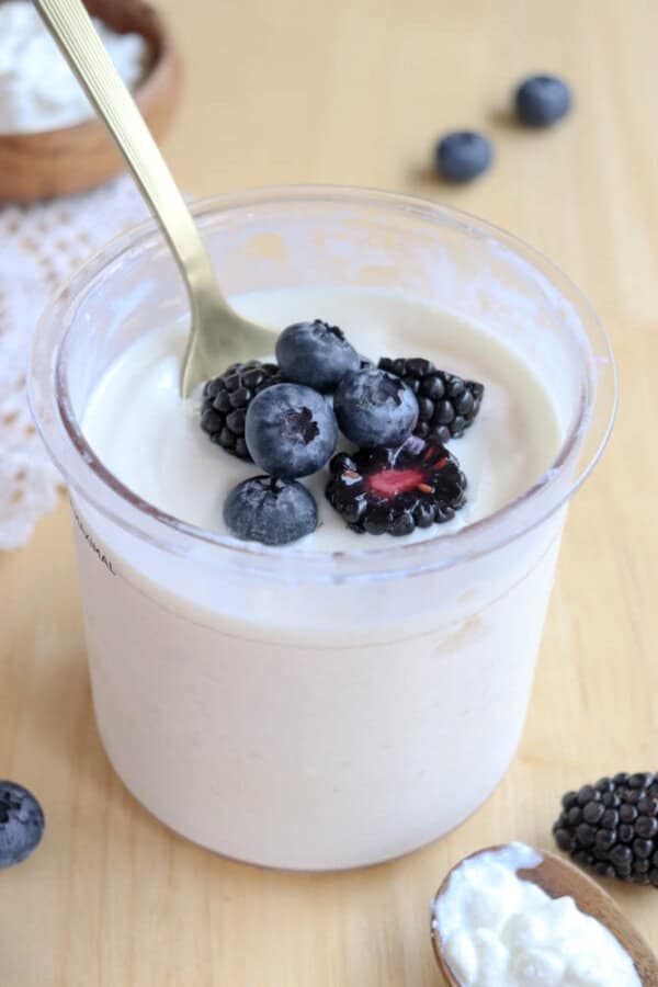 A clear plastic container of cottage cheese ice cream topped with fresh blueberries and blackberries, placed on a light wooden surface. A gold spoon is in the yogurt, and theres a small wooden bowl with more blueberries in the background.