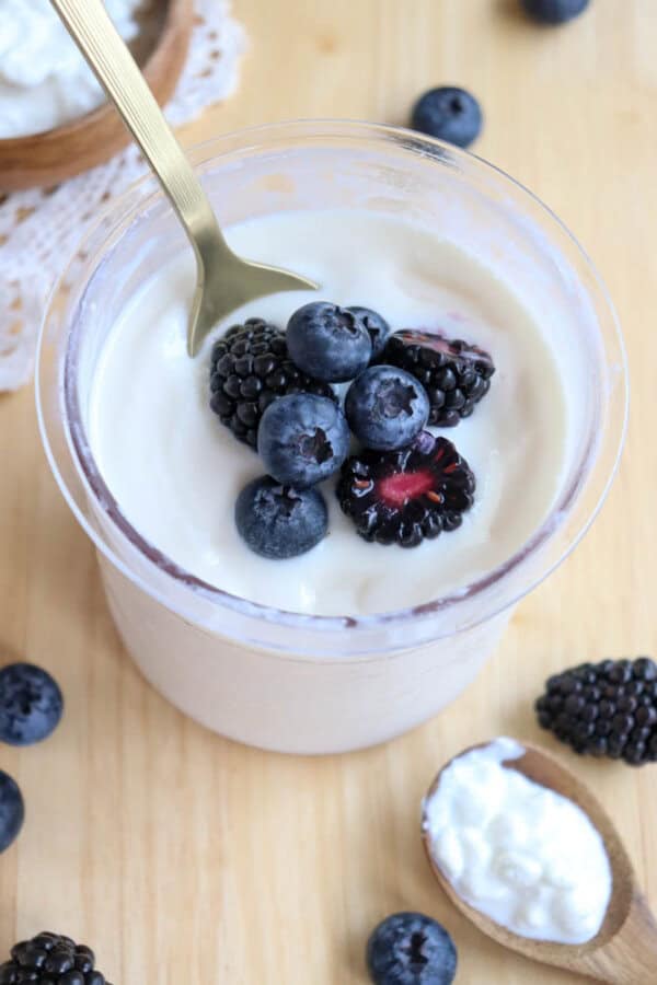 A plastic pint filled with creamy cottage cheese ice cream topped with fresh blueberries and blackberries. A gold spoon rests inside the glass. Additional berries and a wooden spoon with yogurt are scattered on the wooden surface around it.