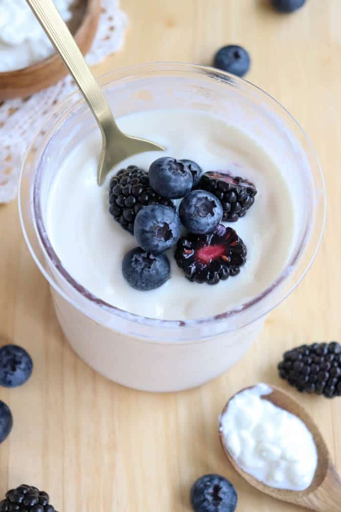 A plastic pint filled with creamy cottage cheese ice cream topped with fresh blueberries and blackberries. A gold spoon rests inside the glass. Additional berries and a wooden spoon with yogurt are scattered on the wooden surface around it.