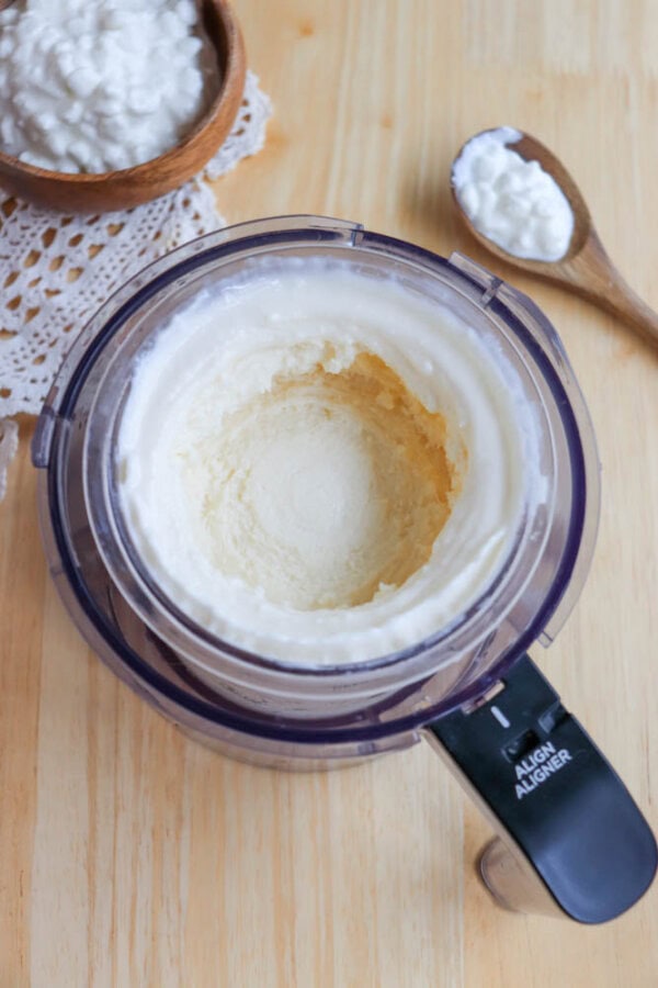 A ninja creami cup on a wooden table filled with creamy mixture. A wooden bowl with clotted cream and a spoon filled with clotted cream are beside it on a lace doily. The processor lid features a black alignment guide.