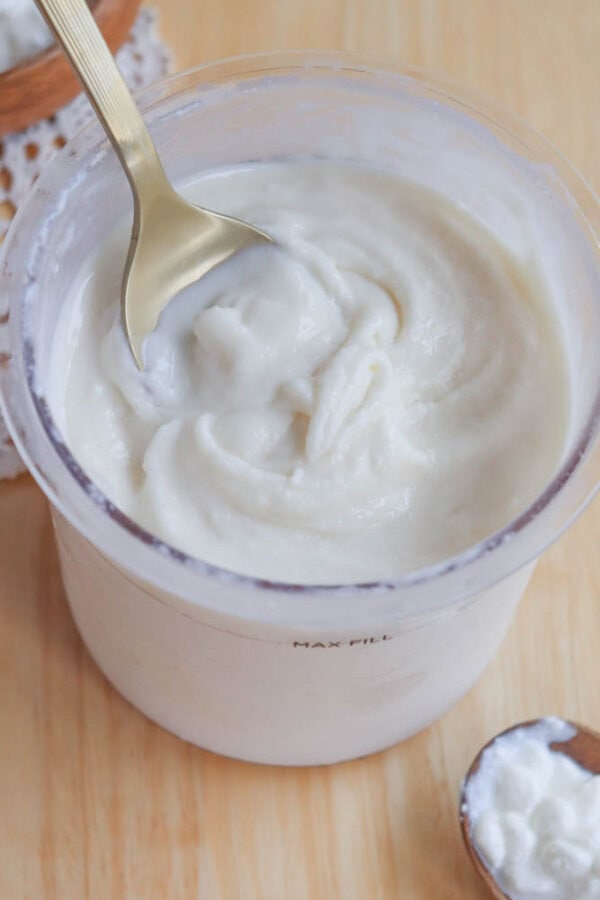 A gold spoon is dipped into a container of cottage cheese ice cream placed on a wooden surface. Theres a small wooden bowl with some yogurt in the foreground. The container has a MAX FILL mark.