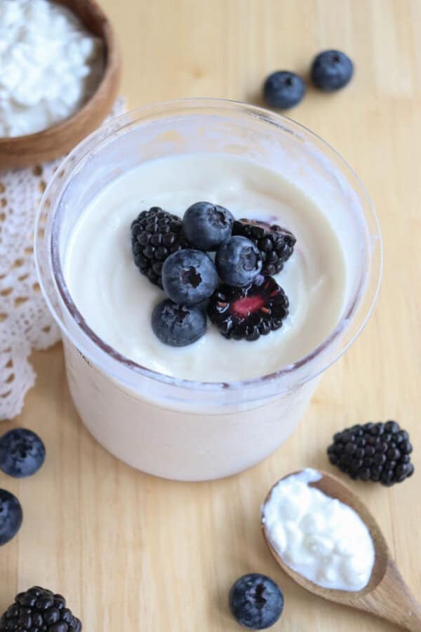 A glass of creamy cottage cheese ice cream topped with fresh blueberries and blackberries sits on a wooden surface. A wooden spoon with yogurt and a few scattered berries and a small bowl with cottage cheese are nearby. A lace cloth is partially visible on the side.