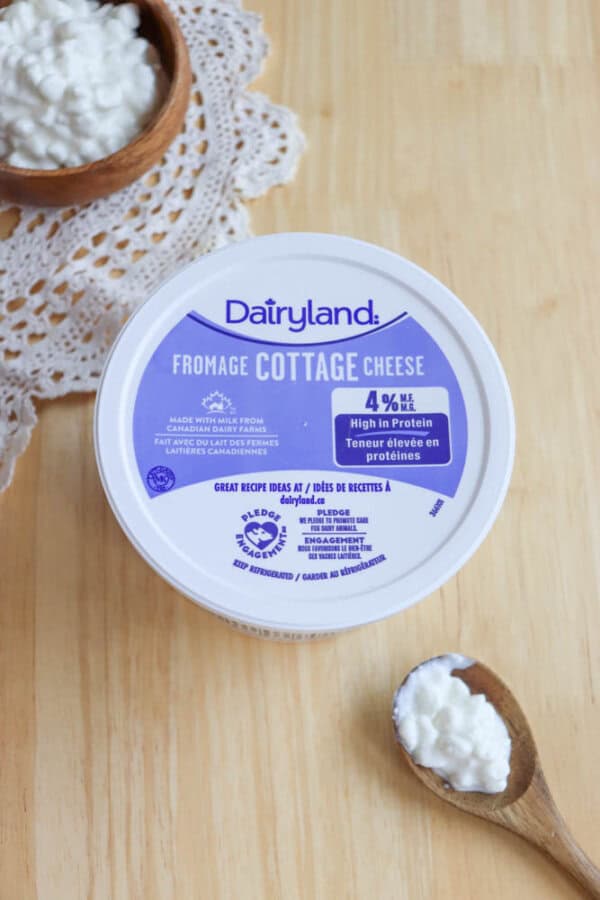 A container of Dairyland cottage cheese sits on a wooden surface. The lid displays text about its protein content. A wooden bowl holds some cottage cheese in the background, with a wooden spoon containing more cottage cheese nearby.