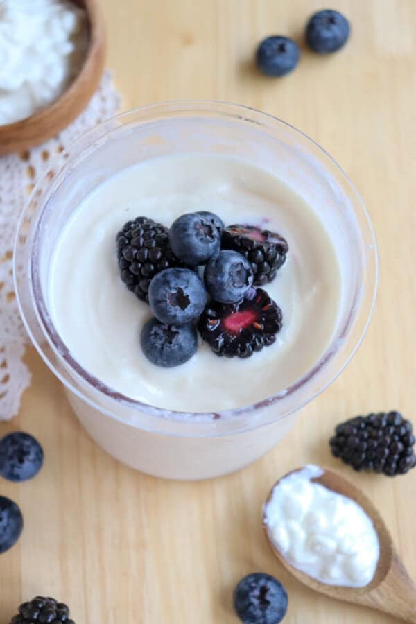 A glass of cottage cheese ice cream topped with fresh blackberries and blueberries sits on a wooden surface. A spoonful of cottage cheese is nearby, along with scattered berries and a small bowl in the background.