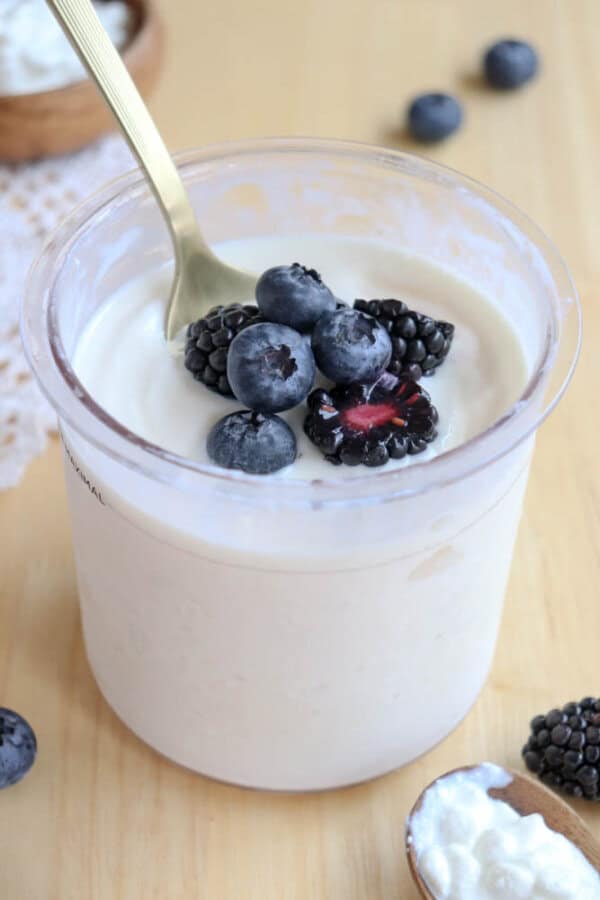 A clear container filled with creamy cottage cheese ice cream  topped with fresh blueberries and blackberries. A gold spoon is dipped into the yogurt. More berries and a small bowl of cottage cheese are in the background on a wooden surface.