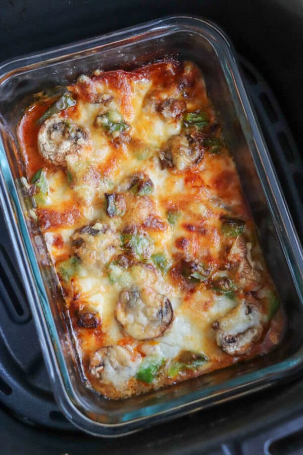 A deliciously baked cottage cheese dish with melted cheese, mushrooms, and green peppers in a clear rectangular glass container. Placed in a black air fryer basket, the top layer is golden-brown with bubbling cheese reminiscent of pizza bowls.