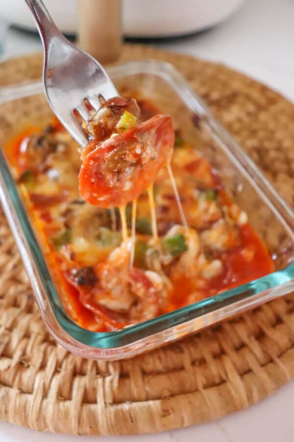 A fork lifts a slice of melty, cheesy pizza from a glass baking dish filled with pizza casserole, enriched with cottage cheese, pepperoni, and vibrant vegetables. The dish rests on a woven placemat.