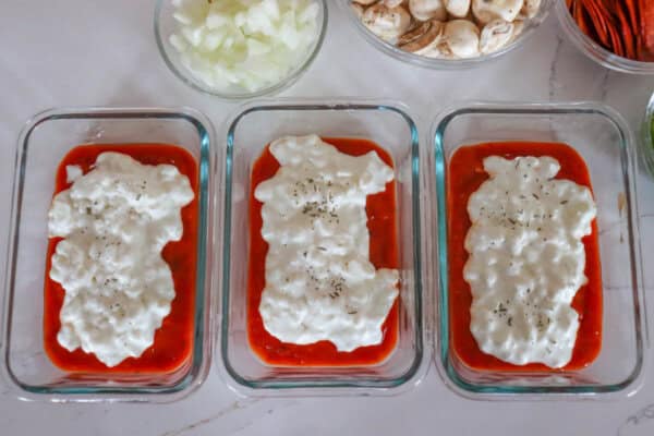 Three rectangular glass containers brim with red tomato sauce, crowned with cottage cheese and herbs. Nearby bowls hold sliced onions and mushrooms, ready to enhance flavor like toppings on a cottage cheese pizza. All are artfully arranged on a pristine white countertop.