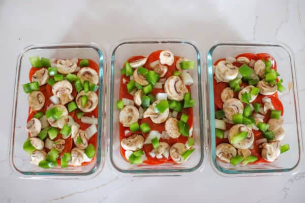 Three glass containers are layered with uncooked ingredients like sliced mushrooms, chopped green bell peppers, and onions on a red sauce base, resembling mini pizza bowls or lasagna-ready dishes, all displayed neatly on a white countertop.