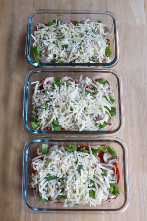 Three glass baking dishes filled with uncooked layered pizza ingredients, including shredded mozzarella cheese, sliced mushrooms, cottage cheese, green bell peppers, and pepperoni, arranged on a wooden table like delicious pizza bowls ready to be baked.